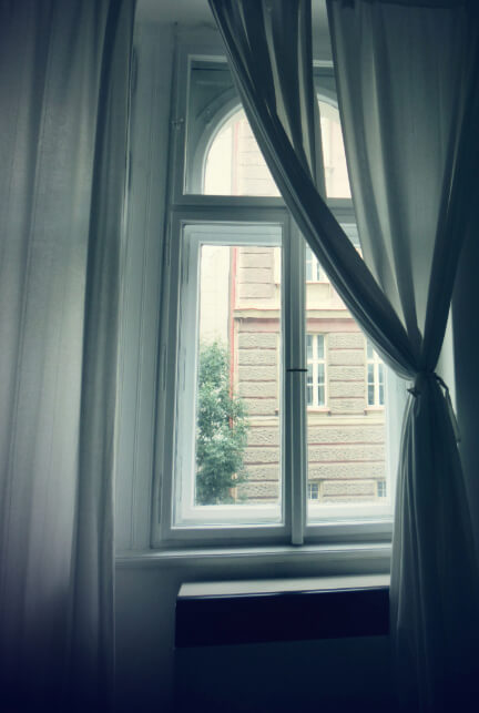 A photograph of a window from inside a room. White soft curtains drape the window and through the glass is a building across the street.